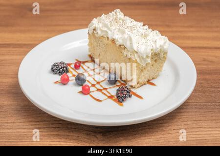 Tranche de gâteau éponge garnie de crème fouettée et de copeaux de chocolat, servie avec des fruits rouges et une sauce au caramel dans une assiette blanche. Banque D'Images