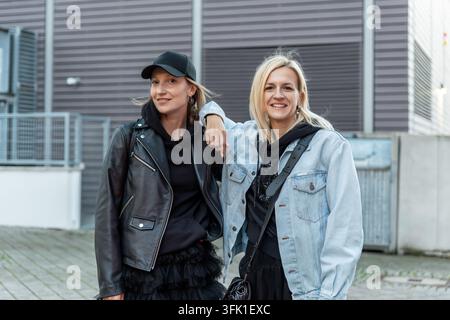 Deux jeunes femmes adultes élégantes d'âge moyen posent ensemble en plein air dans un style de mode urbain, souriant avec une confiance détendue. Portrait amical capturant l'amitié Banque D'Images