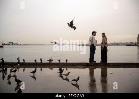 Un jeune couple se tient ensemble sur le front de mer à Kadıköy, Istanbul, Turquie, le 16 septembre 2026, entouré de pigeons sur une promenade mouillée. Banque D'Images