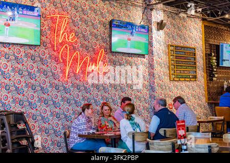 Arkansas, États-Unis, 07 avr. 2024 – Des amis se rassemblent autour d'une table en bois dans un restaurant vibrant doté d'un papier peint à motifs et d'une néon lumineuse Banque D'Images