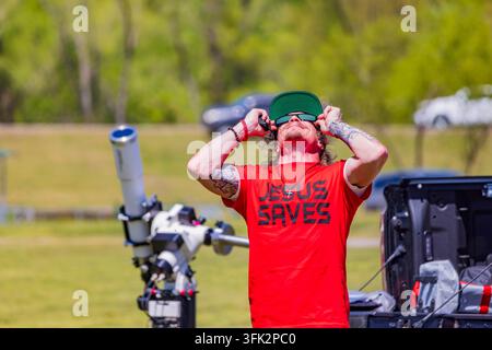 Arkansas, États-Unis, 08 avr. 2024 – Un homme portant un t-shirt rouge avec du texte se tient en plein air tout en ajustant ses lunettes de soleil sous le soleil éclatant. Banque D'Images