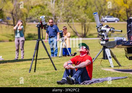Arkansas, États-Unis, 08 avr. 2024 – Un homme en chemise rouge est assis sur l'herbe tandis que d'autres utilisent des télescopes pour des observations astronomiques par une journée ensoleillée Banque D'Images