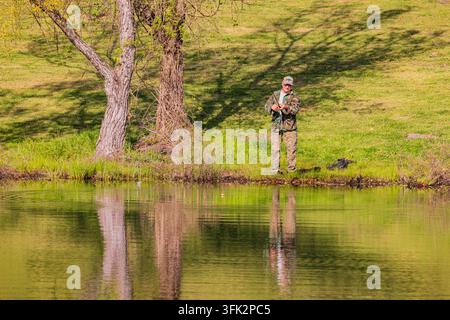 Arkansas, États-Unis, 08 avr. 2024 - Un homme vêtu de vêtements de camouflage se tient sur la berge herbeuse d'un lac calme à l'ombre de grands arbres. Banque D'Images