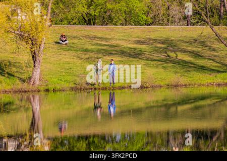 Arkansas, États-Unis, 08 AVR 2024 - Deux personnes se tiennent au bord de la rivière herbeuse avec des cannes à pêche tandis qu'une autre personne est assise sur la colline dans le Banque D'Images