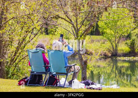 Arkansas, États-Unis, 08 AVR 2024 – Deux personnes sont assises sur des chaises de camping turquoise au bord d'un lac calme tandis qu'une personne prend une photo du paysage printanier pittoresque Banque D'Images