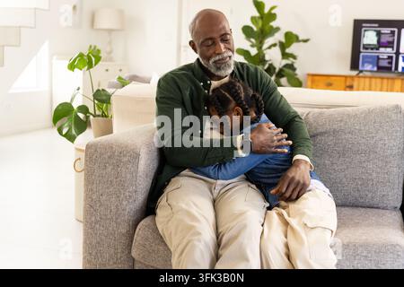 Homme afro-américain mûr tenant une petite fille blottie sur un canapé dans un cardigan avec une montre connectée Banque D'Images