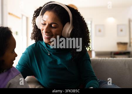 Mère et fille afro-américaines assises sur le canapé du salon, portant des écouteurs blancs Banque D'Images