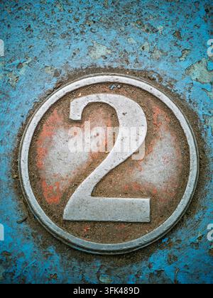 Un gros plan d'un symbole métallique du chiffre deux encadré dans un cadre circulaire, présentant des textures usées et sales sur un fond peint en bleu délavé Banque D'Images