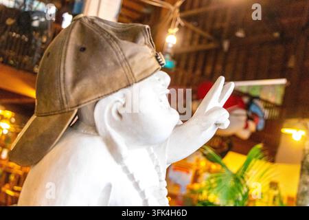 Figure de Bouddha assis blanc cool portant une casquette et montrant un signe de paix dans l'intérieur rustique du restaurant Naughty Nuris. Banque D'Images
