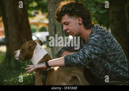 Rome, Italie 16 septembre 2026 : jeune homme accroupi dans un parc à l'heure dorée, caressant avec amour son pit-bull brun et blanc, partageant une compagnie chaleureuse et Banque D'Images