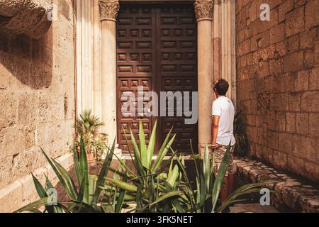 Cagliari, Italie 16 septembre 2026 : homme debout devant une porte en bois historique ornée encastrée dans des colonnes de pierre, regardant vers le haut des panneaux sculptés par une journée ensoleillée da Banque D'Images