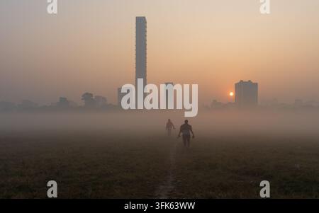 Vue de deux personnes marchant à travers un champ herbeux dans la brume matinale, avec le panorama urbain et le lever du soleil en arrière-plan à Kolkata, au Bengale occidental Banque D'Images