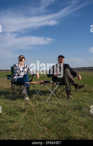 Couple profitant d'un moment de pique-nique relaxant, assis sur des chaises de camping à côté d'une tente, buvant ensemble de la bière dans un vaste champ vert sous un ciel bleu clair Banque D'Images