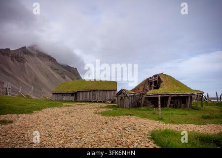 Des cabanes en bois érodées se dressent sur une prairie montagneuse verte sous des pentes rocheuses escarpées et des nuages bas et brumeux dans un paysage rural isolé. Banque D'Images