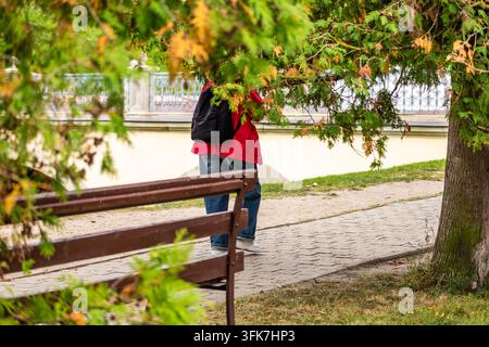 Person portant une veste rouge, un jean bleu et un sac à dos sombre marche le long d'un sentier pavé du parc encadré de branches vertes et de feuilles d'automne. Un banc de parc en bois Banque D'Images