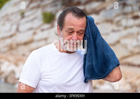 Un homme d'âge moyen en T-shirt blanc sèche sa tête avec une serviette bleue en plein air. Banque D'Images