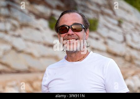 Un homme souriant d'âge moyen portant des lunettes de soleil se tient à l'extérieur devant un fond rocheux. Banque D'Images