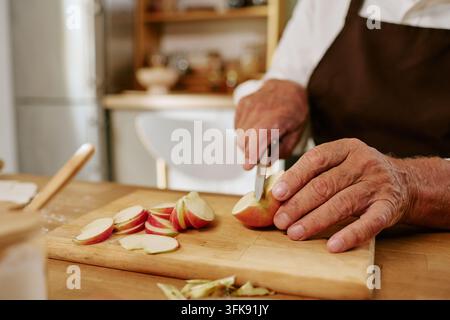 Homme âgé coupant des pommes rouges fraîches sur une planche en bois dans la cuisine Banque D'Images