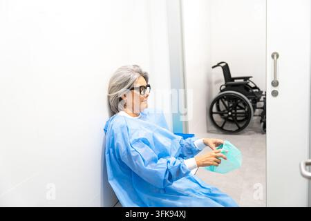 Dame âgée en robe bleue tenant un bassin tout en s'appuyant contre un mur Banque D'Images
