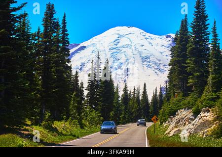 Une route sinueuse traverse une forêt luxuriante à feuillage persistant en direction du sommet enneigé du mont Rainier dans la région de Sunrise de Washington. Des voitures empruntent la route Banque D'Images