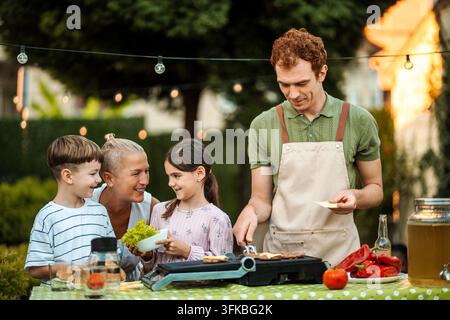 Famille profitant d'un barbecue dans le jardin en cuisinant des burgers ensemble Banque D'Images