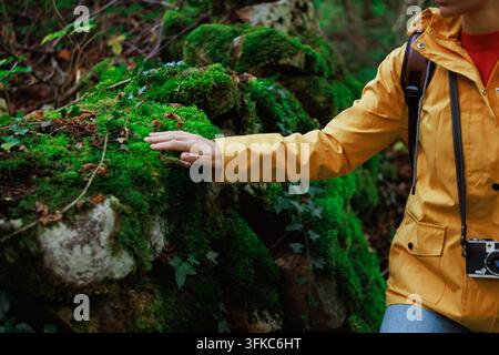 Femme en imperméable jaune et sac à dos touche doucement une mousse verte vibrante sur un gros rocher dans une forêt paisible, ressentant une connexion calme avec la nature Banque D'Images