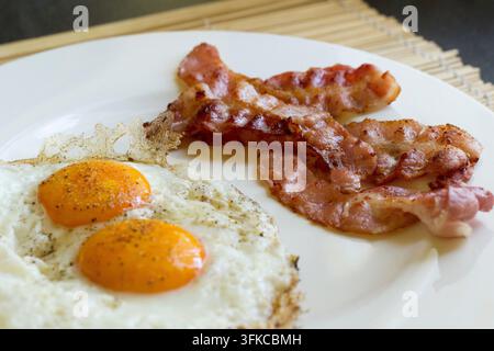 Close-up de deux œufs au plat avec poivre noir et lanières de bacon croustillantes sur une assiette blanche, tapis en bambou en arrière-plan Banque D'Images