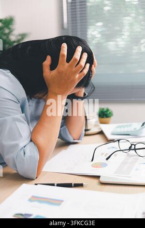 Femme d'affaires submergée tenant sa tête à un bureau avec ordinateur portable, calculatrice et rapports, montrant un burn-out, un stress au travail, une pression des délais, une w financière Banque D'Images