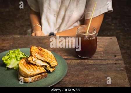 Femme dégustant un panini au poulet grillé avec un verre d'Americano glacé sur une table en bois Banque D'Images