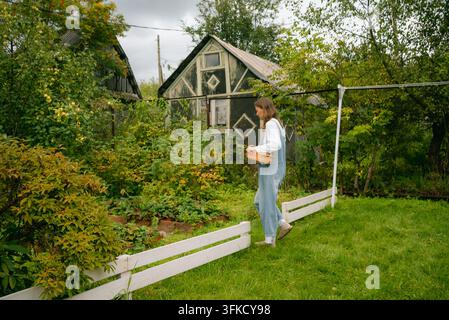 Jeune agricultrice portant une salopette en jean marche le long d'une clôture blanche en bois portant un panier en osier dans un jardin luxuriant et envahi par la végétation à côté d'une petite maison Banque D'Images