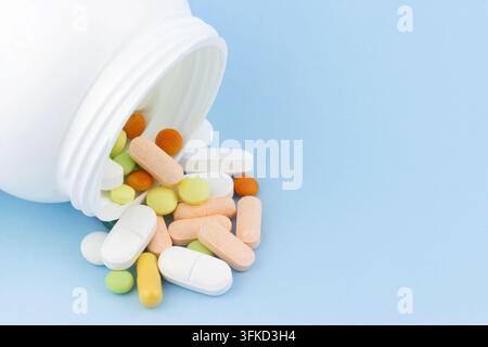 Une bouteille de médicament en plastique blanc renversée déversant des pilules, des comprimés et des capsules multicolores sur un fond bleu. Banque D'Images