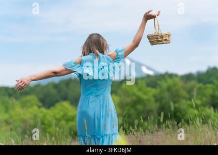 Vue arrière d'une femme en robe à pois bleus profitant de l'été en plein air, les bras écartés tenant un panier en osier. Bras féminins grands ouverts dans une pose joyeuse Banque D'Images
