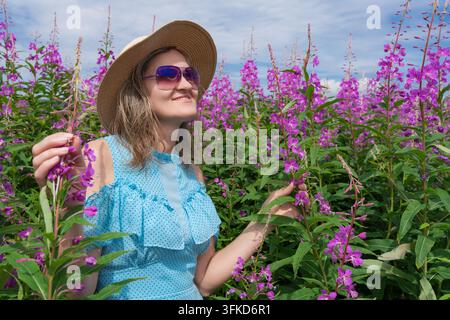 Femme en lunettes de soleil, robe à pois bleus et chapeau de paille souriant parmi de grandes fleurs sauvages violettes estivales. Femme joyeuse touchant doucement une fleur violette vibrante Banque D'Images