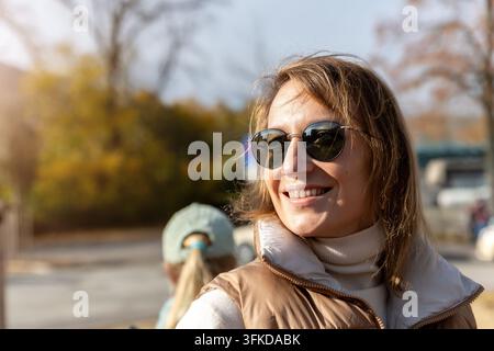 Femme souriante portant des lunettes de soleil profitant de la lumière chaude de l'automne en plein air dans un portrait de style de vie naturel. Un feuillage doré et un fond urbain doux créent Banque D'Images