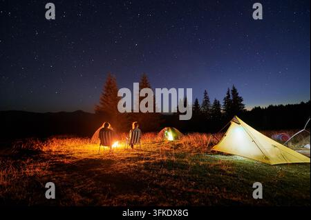 Deux campeurs assis autour d’un feu de camp sous un ciel étoilé, entourés de tentes illuminées. Une nuit sereine et claire dans les montagnes crée une atmosphère paisible, idéale pour l’observation des étoiles et la détente dans la nature. Banque D'Images