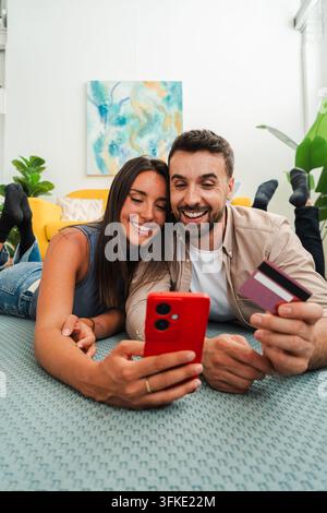 Un couple joyeux est allongé sur un lit et partage un moment ludique en utilisant un smartphone rouge vif et une carte de crédit comme accessoires. La scène capture l'affection Banque D'Images