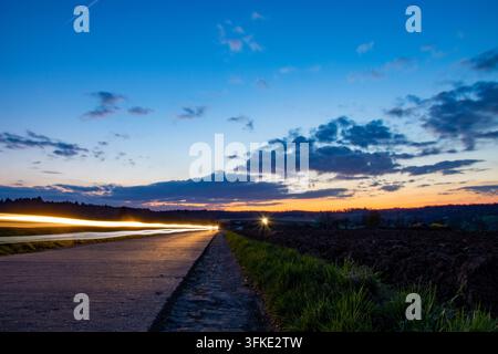 Photographie à longue exposition des traînées de phares vives sur une route de campagne déserte traversant des champs pendant le crépuscule, avec un coucher de soleil dramatique bleu et orange Banque D'Images