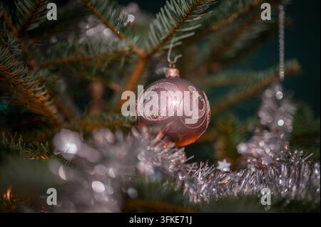 Un plan rapproché, artistique, d'une boule de Noël rose décorative avec des motifs de tourbillons pailletés suspendue à un pin vert luxuriant, entourée d'argent t Banque D'Images