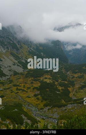 Vue panoramique d'une vallée de haute montagne avec un lac alpin sombre entouré de pentes rocheuses, d'une végétation basse et de nuages bas. Banque D'Images