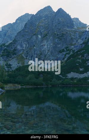 Un lac de montagne serein reflète des pics rocheux imposants et des forêts de pins verts sous un ciel matinal doux. Banque D'Images