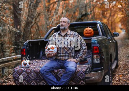 Homme barbu tenant un citrouille peinte en crâne près d'un pick-up, idéal pour la publicité Halloween et les campagnes saisonnières en extérieur. Banque D'Images