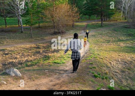 Soignant marchant derrière deux enfants sur un sentier de terre à travers une forêt de printemps avec des pins et des bouleaux, une végétation variée et une clairière herbeuse ouverte Banque D'Images