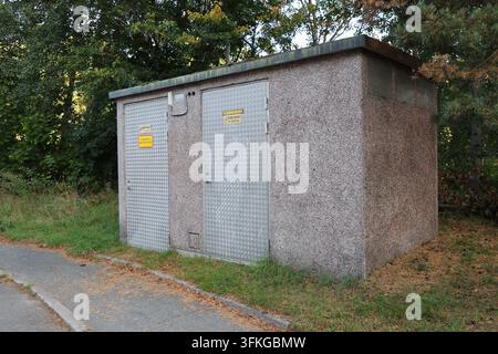 Un bâtiment de sous-station électrique en béton avec une porte en plaque métallique à motifs comportant des panneaux d'avertissement, situé près d'arbres verts. Banque D'Images