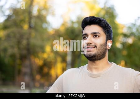 Jeune homme souriant portant des écouteurs sans fil profitant de contenus audio en se détendant dans un parc, représentant le style de vie, la technologie et la connexion avec la nature Banque D'Images