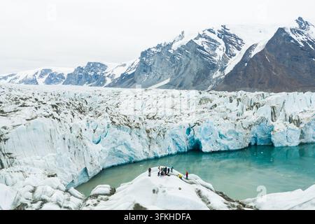 Vue aérienne d'un glacier massif avec des formations de glace bleu profond et des personnes debout sur un affleurement rocheux près d'un lac glaciaire turquoise, entouré de neige Banque D'Images