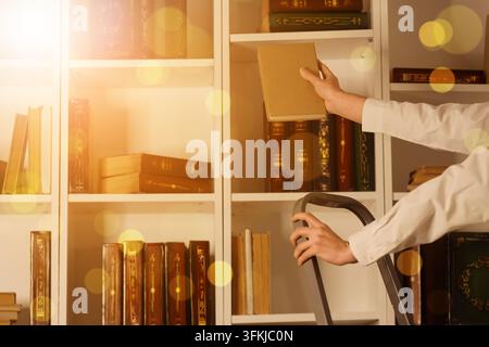 Éducation et recherche, personne atteignant un livre sur une étagère élevée dans une bibliothèque avec une échelle pour trouver des connaissances et des ressources académiques pour l'apprentissage Banque D'Images