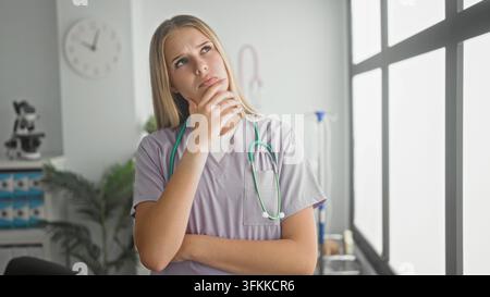 Jeune femme médecin blonde portant une blouse lilas avec stéthoscope, main sur le menton, réfléchissant à un dossier patient près de la fenêtre dans une clinique ; inquiète. Banque D'Images