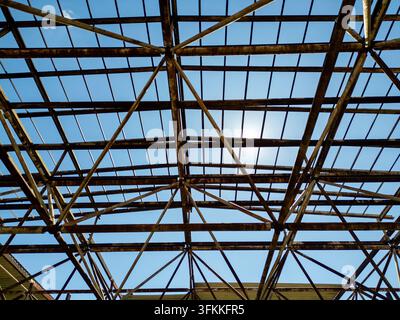 Vue de bas en haut d'une charpente de toit métallique rouillée et d'une structure de cadre d'un entrepôt industriel ou hangar abandonné sous un ciel ensoleillé brillant avec éblouissement du soleil Banque D'Images