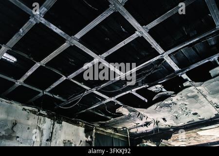Une pièce intérieure montre un plafond fortement endommagé par le feu avec une grille métallique exposée, des panneaux manquants et des traces de carbonisation et de fumée visibles, destruction Banque D'Images