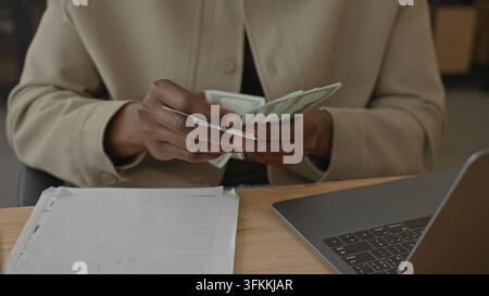 Homme comptant des dollars avec ses mains au-dessus d'un ordinateur portable et de papiers sur un bureau à l'intérieur d'un bâtiment ; concentration sur la gestion de l'argent. Banque D'Images